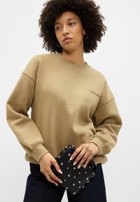 Khaki oversize mikina s volnými rameny, žebrovanými manžetami a vyšitým textem. Černá texturovaná taška se stříbrnými hroty. Tmavé džíny dotvářejí outfit.