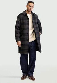 Manteau à carreaux noir et gris avec un devant à boutons, deux poches latérales et un col, porté sur un pull beige et un jean foncé. Des chaussures marron complètent la tenue.