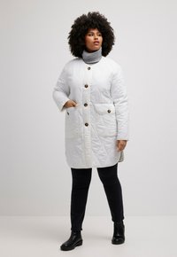 Bílý prošívaný kabát s velkými předními kapsami a hnědými knoflíky. Nositý přes šedý rolák a v kombinaci s černými přiléhavými kalhotami a kotníkovými botami.