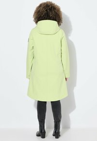 Manteau vert clair avec une capuche, présentant une coupe droite et des poches latérales, associé à un pantalon noir ajusté et des bottines.