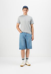 Giovane uomo che indossa una maglietta grigia, pantaloni corti di jeans, sneakers bianche, calzini a righe e un cappellino di jeans, in piedi contro uno sfondo bianco.