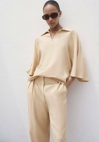 Beige übergroße Bluse mit Kragen, kombiniert mit beige gefalteten Hosen. Glattes Material, lockere Passform und dezente Drapierungsdetails. Braune Sonnenbrille.