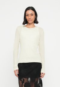 Γυναίκα με σκούρα μαλλιά μέχρι τους ώμους φοράει λευκή μπλούζα με μακριά μανίκια και μαύρη δαντελένια φούστα, στέκεται μπροστά σε απλό φωτεινό φόν το.