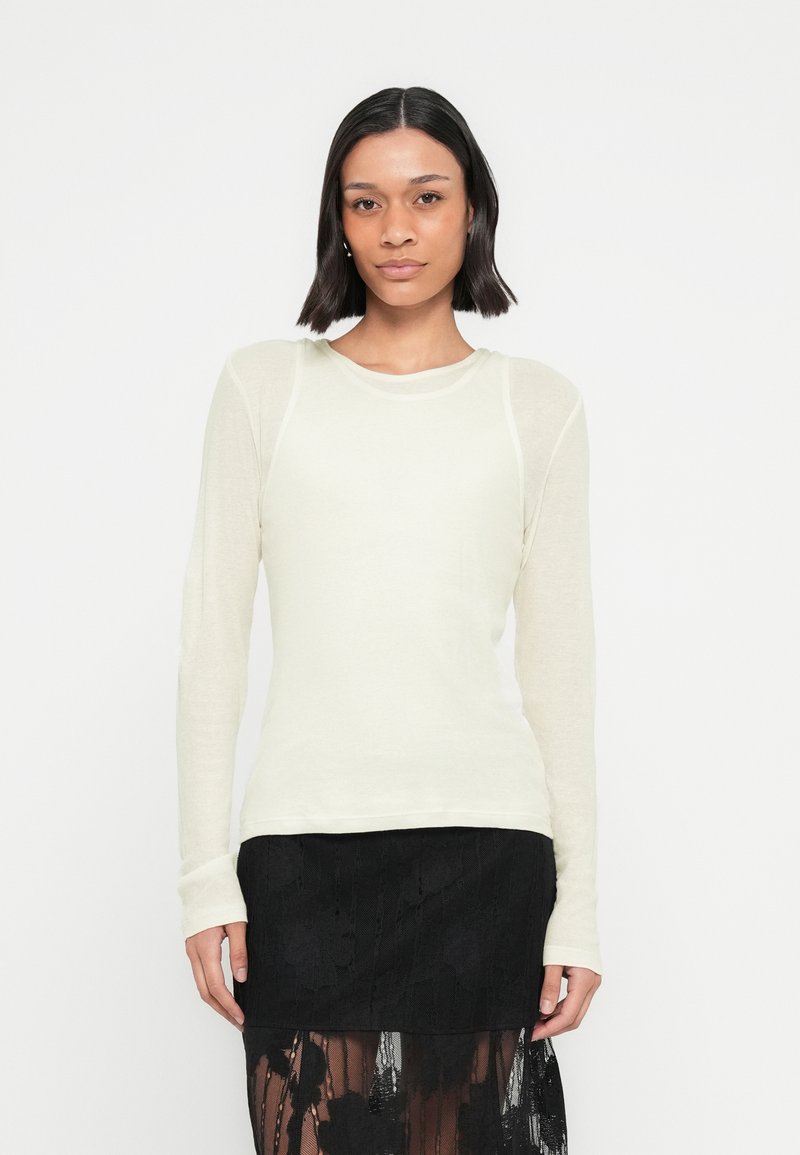 Γυναίκα με σκούρα μαλλιά μέχρι τους ώμους φοράει λευκή μπλούζα με μακριά μανίκια και μαύρη δαντελένια φούστα, στέκετ αι μπροστά σε απλό φωτεινό φόντο.