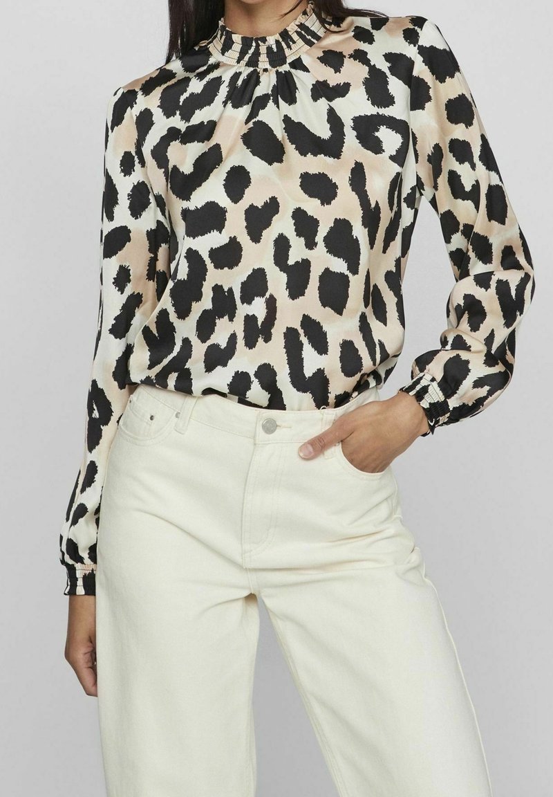 Blouse à imprimé léopard avec un col haut à volants, des manches longues et des poignets élastiques. Portée avec un jean blanc taille haute. Texture de tissu lisse.