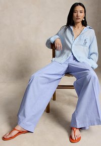 Γυναίκα καθισμένη σε ξύλινη καρέκλα φορώντας ανοιχτό γαλάζιο φαρδύ πουκάμισο, φαρδιά λεβαντίνια παντελόνια και πορτοκαλί σαγιονάρες μπροστά σε μπεζ φόντο.