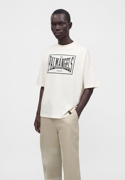 Άνδρας μοντέλο που φοράει λευκό oversized T-shirt με κείμενο "L.A. Palm Angels" και μπεζ παντελόνι, στέκεται μπροστά σε απλό ανοιχτό γκρι φόντο.