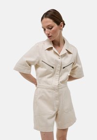 Jumpsuit beige à manches courtes avec un col, comportant deux poches zippées sur la poitrine et des poches latérales. Fabriqué en tissu solide.