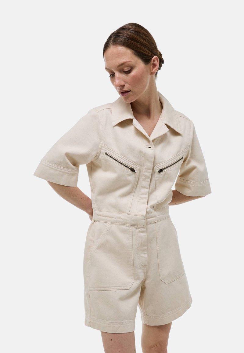 Jumpsuit beige à manches courtes avec un col, comportant deux poches zippées sur la poitrine et des poches latérales. Fabriqué en tissu solide.