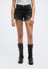 Γυναίκα που φοράει μαύρα κομμένα τζιν σορτς, λευκό μπλουζάκι και μαύρες δερμάτινες μπότες μέχρι το γόνατο, στέκεται μπροστά σε ανοιχτό γκρι φόντο.