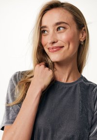 Grijze fluwelen t-shirt met korte mouwen, ronde halslijn en subtiele glans. Model raakt haar aan en draagt kleine gouden ringoorbellen. Glad van structuur.