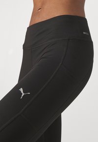 Leggings deportivos negros hechos de material que evacua la humedad, con una cinturilla alta y un logo en contraste en el muslo. Textura suave y detalles de costura.
