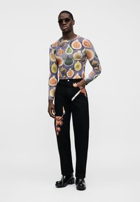 Úzky top s farebnými potlačami fig a čierne nohavice s potlačou rúk držiacich štetec a nôž, nosené s čiernymi topánkami a slnečnými okuliarmi.