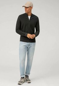 Schwarzer Pullover mit Reißverschluss aus leichtem Strick, kombiniert mit hellblauen, legeren Jeans und einer beigen Mütze. Sneakers mit grauen Akzenten vervollständigen den Look.