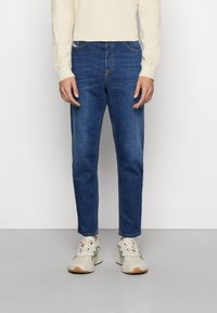 Blaue Denim-Jeans mit schmaler Passform, featuring gestickte Details, Fünf-Taschen-Design und mittlere Waschung, getragen mit beigen Sportschuhen.