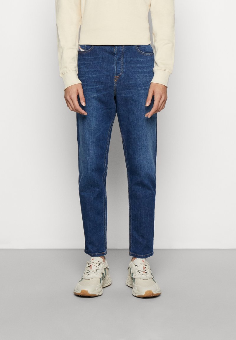 Modré džíny z denimového materiálu se slim fit střihem, s prošívanými detaily, pětibesedovým designem a středním praním, nošené s béžovými sportovními botami.