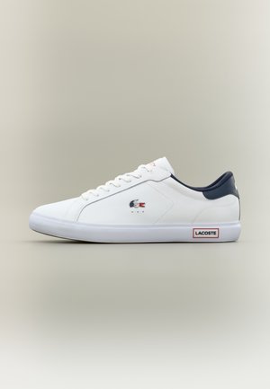 Valge Lacoste madala profiiliga toss, millel on tumesinine krae, valged nöörid, krokodilli logo küljel ja Lacoste silt valgel kummitaldal.