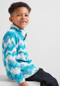 Fleece pullover in turquoise and white, featuring a zigzag pattern, a high collar, and a front zipper. Worn with dark pants.