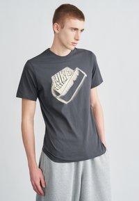 Sivé bavlnené tričko s okrúhlym výstrihom a grafickou potlačou loga Nike v kontrastnej svetlej farbe. V kombinácii so sivými nohavicami.