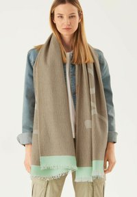 Écharpe marron avec un motif texturé, dotée d'une bordure à franges vert menthe. Portée avec une veste en denim sur un haut léger.