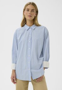 Chemise boutonnée bleu clair avec des rayures verticales en bleu foncé et orange. Elle présente un col à pointes écartées et des poignets blancs. Tissu doux.