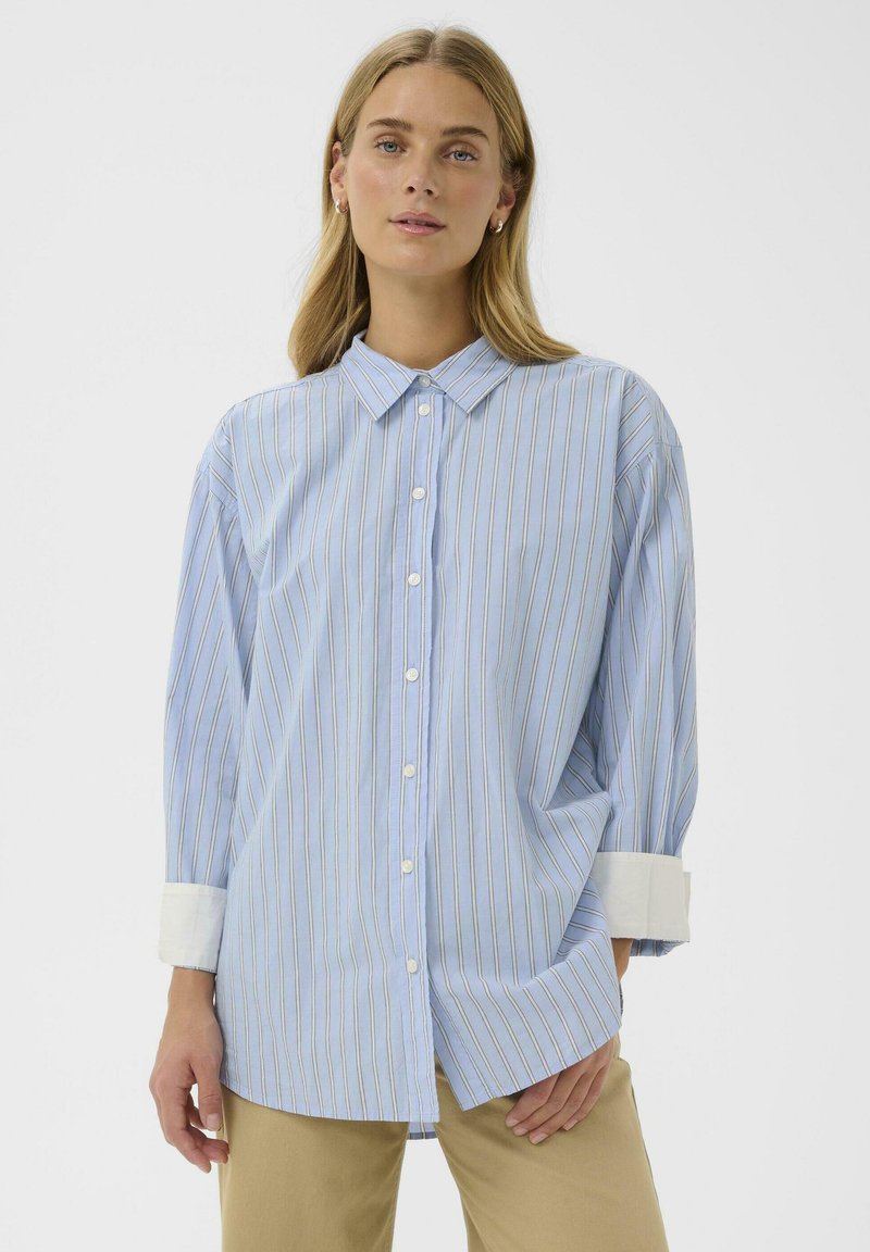 Chemise boutonnée bleu clair avec des rayures verticales en bleu foncé et orange. Elle présente un col à pointes écartées et des poignets blancs. Tissu doux.