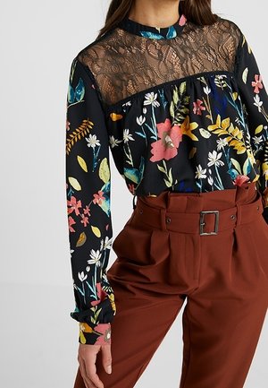 Musta kukkakuvioinen paita, jossa on läpinäkyvä pitsiyläosa, yhdistettynä korkeavyötäröisiin ruosteenvärisiin housuihin, joissa on leveä vyö ja solki.