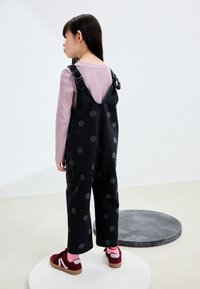 Salopette en denim noir à pois gris, avec bretelles ajustables et poches latérales, assortie à un t-shirt à manches longues rose clair et des chaussures bordeaux.