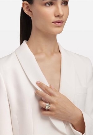 Silber- und Goldring mit gravierten Mustern, getragen an einer Hand vor einem hellen Blazer mit breitem Kragen.
