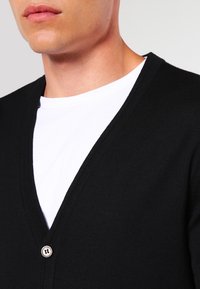 Cardigan nero con design a V, dotato di chiusura con un singolo bottone e tessuto testurizzato. Indossato sopra una maglietta bianca.