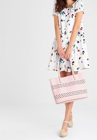 Roze draagtas met geperforeerde horizontale strepen en contrasterende stippen, gemaakt van glad faux leer. De tas heeft stevige handvatten en een gestructureerde vorm.