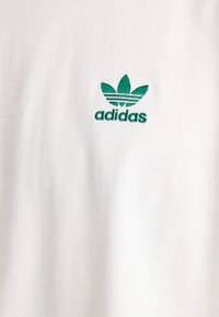 Tricou alb din bumbac, cu logo Adidas trefoil brodat în verde pe partea stângă a pieptului, cu o textură netedă și un design clasic.