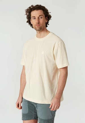 Camiseta de algodón color crema con un corte relajado, mangas cortas, cuello redondo y un pequeño detalle de logo en el pecho. Textura suave.