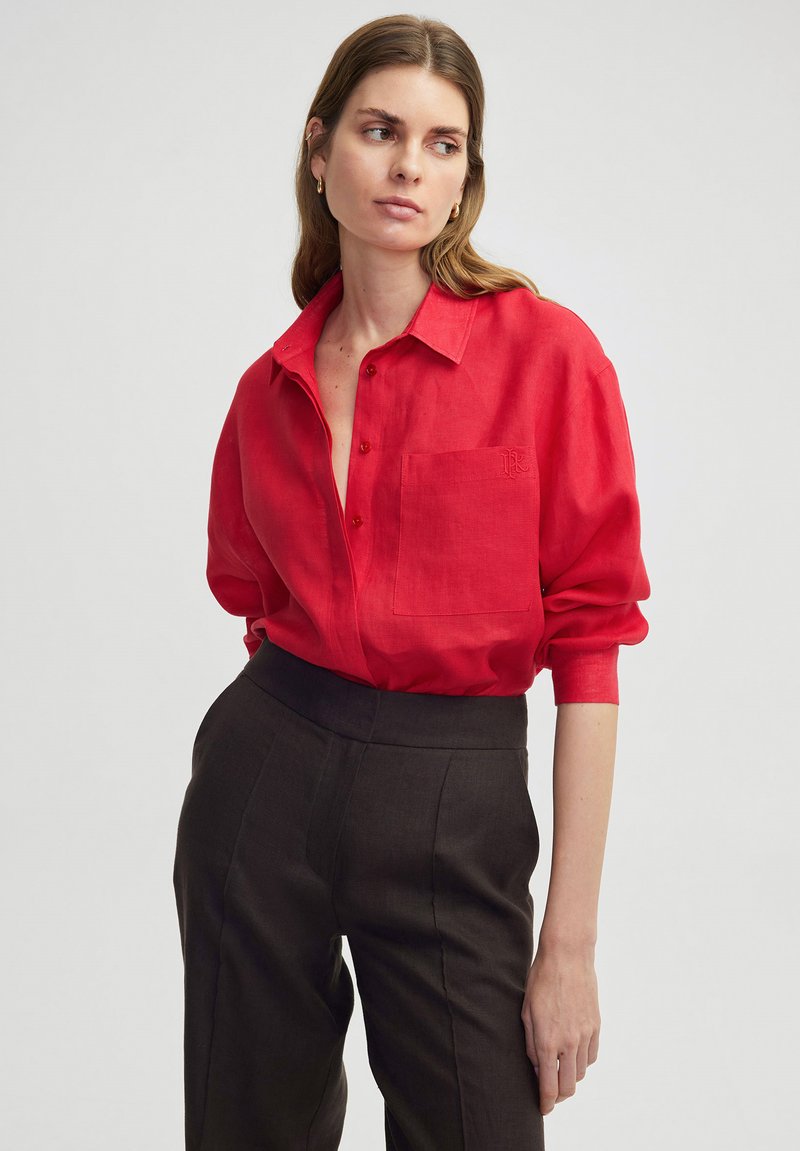 Chemise en lin rouge avec un col, une fermeture à boutons sur le devant et une poche sur la poitrine. Les manches longues se rassemblent aux poignets. Portée avec un pantalon sombre taille haute.
