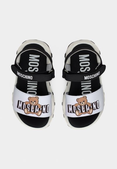 Μαύρα και άσπρα σανδάλια Moschino με γραφικό αρκουδάκι στο μπροστινό λουράκι και το όνομα της μάρκας στα λουράκια και την υποδήλωση, από ψηλά.