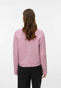 Rožinės spalvos tekstūruotas mezgimo megztinis su deimanto raštu, banguotu apačios kraštu ir ilgomis rankovėmis, dėvimas ant juodų kelnių.