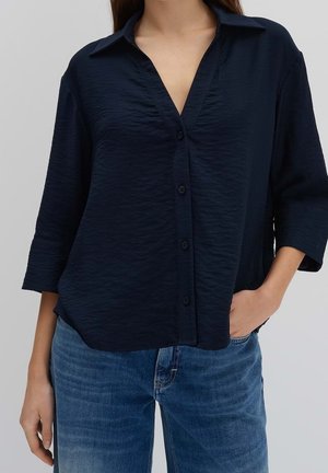 Mujer vestida con una blusa azul oscuro abotonada con cuello y mangas 3/4, combinada con jeans azules, con la mano reposando en el bolsillo.