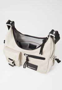 Beige nylon kézitáska fekete kiegészítőkkel, cipzáras rekeszekkel, állítható pánttal és elülső zsebből. Simább textúra minimális márkázással.