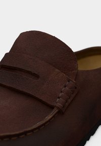 Καφέ μοκασίνι από σουέντ με λουράκι στο πάνω μέρος, λεπτομερείς ραφές στο πλάι και απαλή, υφή στην επιφάνεια.