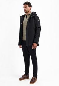 Man in een zwarte hoodie, een grijze sweater met rits tot kwart, zwarte broek en bruine veterschoenen, staande op een witte achtergrond.