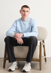Svetlo modra majica, črne kavbojke in sivi tenis čevlji z črnimi detajli. Model sedi na svetlem beigem stolu z naravno leseno konstrukcijo.