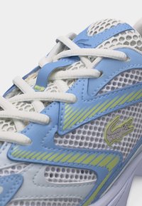 Λευκό και μπλε πλεκτό αθλητικό παπούτσι με λευκά κορδόνια και λογότυπο κροκόδειλου με πράσινες λεπτομέρειες στο πλάι.