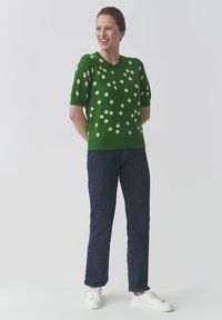 Pull vert à manches courtes avec des pois blancs, associé à un jean en denim foncé et des baskets blanches. Le modèle se tient avec les mains derrière le dos.