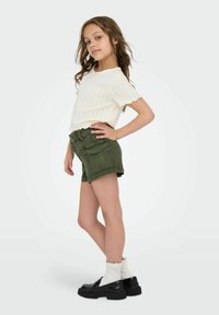 Vitt strukturerat kortärmat linne med scallop-detaljer, parat med gröna cargoshorts och svarta plattformsskor, accentuerat med vita volangstrumpor.