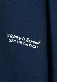 Tmavomodrý textil s bielym vyšívaným textom: "Víťazstvo je podávané HRA, SET, MÁČ." Materiál má hladký, štruktúrovaný povrch.
