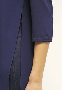 Marinblus med trekvartslång ärm, med sidoflikar och en slät textur. Tyget verkar lätt och något draperat.