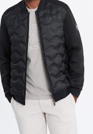 Mann trägt eine schwarze gesteppte Jacke mit Reißverschluss über einem hellgrauen Grafik-T-Shirt und weißen Hosen, die Hände in den Taschen.