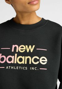 Personne portant un sweat à capuche noir avec le texte "new balance ATHLETICS INC." en douces couleurs pastel sur le devant.