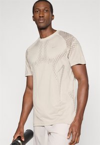 Μπεζ αθλητικό μπλουζάκι με πλέξη για να αναπνέει και αντίθετους φωτεινούς τόνους, διαθέτει στρογγυλή λαιμόκοψη και κοντά μανίκια, συνδυασμένο με μια μαύρη μπουκάλι νερού.