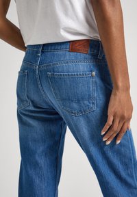 Jeans de mezclilla azul oscuro con una pierna ligeramente ajustada, que presentan bolsillos traseros, un parche de cuero marrón en la cintura y sutiles detalles de costura.